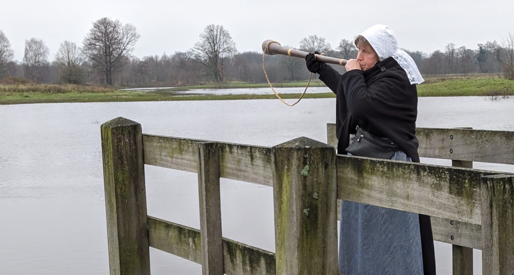 Vrouw in traditionele kleding blaast op een midwinterhoorn op een houten steiger bij een waterlandschap.
