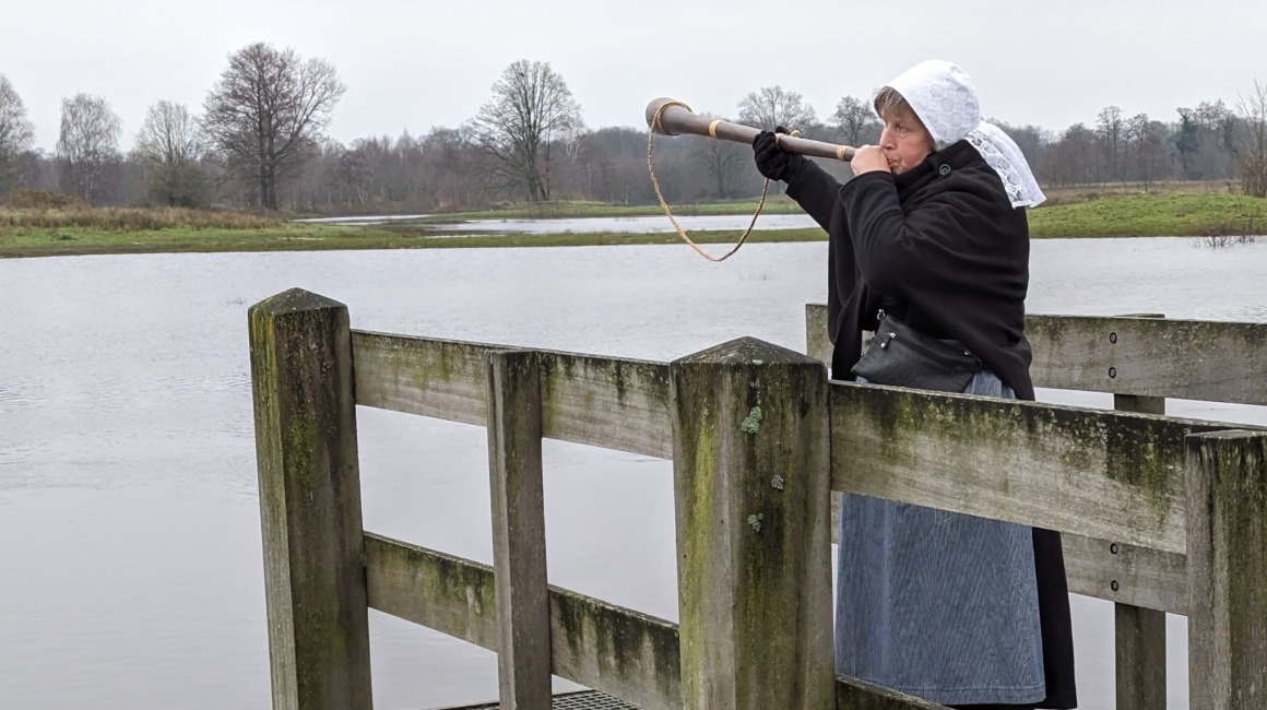 Vrouw in traditionele kleding blaast op een midwinterhoorn op een houten steiger bij een waterlandschap.