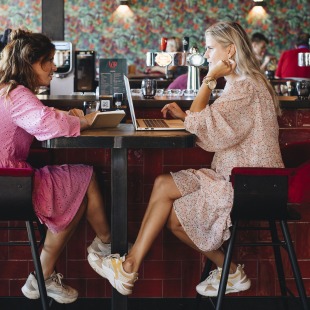 Twee vrouwen in roze jurken zitten tegenover elkaar in een café, werken op een laptop en notitieblok.