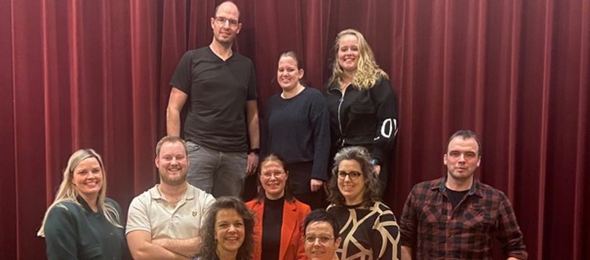 Groepsfoto van tien mensen, sommige zittend op een trap, voor een rode gordijn in een zaal.