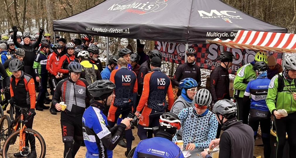 Groep wielrenners met helmen verzamelen bij een tent voor een pauze tijdens een toertocht in het bos.