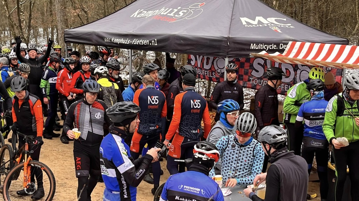 Groep wielrenners met helmen verzamelen bij een tent voor een pauze tijdens een toertocht in het bos.
