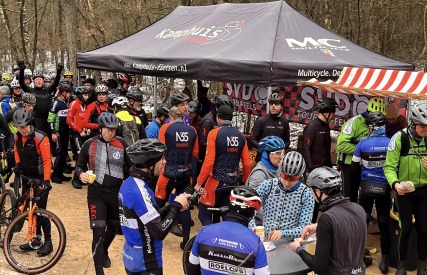 Groep wielrenners met helmen verzamelen bij een tent voor een pauze tijdens een toertocht in het bos.