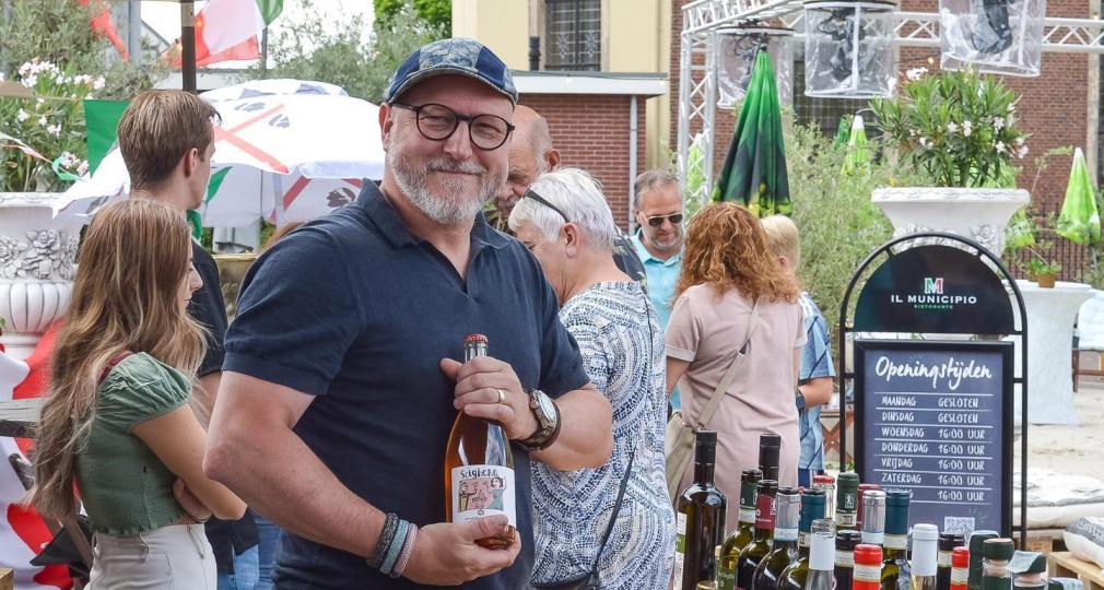Man met fles wijn poseert glimlachend bij een buitentafel vol wijnflessen tijdens een evenement.