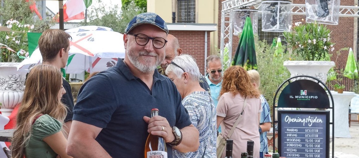 Man met fles wijn poseert glimlachend bij een buitentafel vol wijnflessen tijdens een evenement.