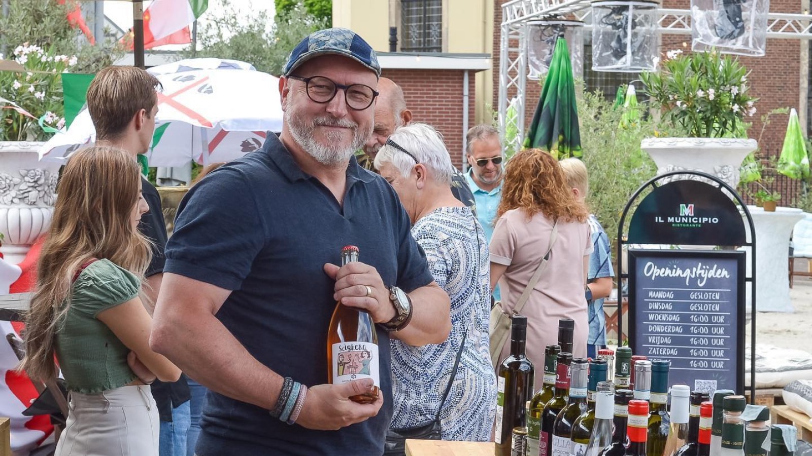 Man met fles wijn poseert glimlachend bij een buitentafel vol wijnflessen tijdens een evenement.