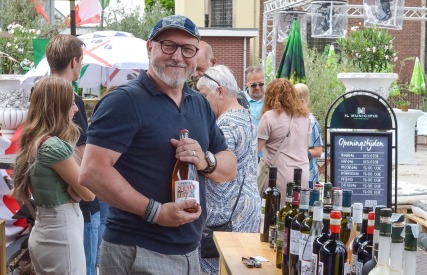 Lächelnder Mann mit Weinflasche steht an einem Tisch mit Weinflaschen auf einer Veranstaltung im Freien.