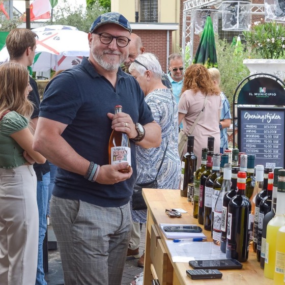 Man met fles wijn poseert glimlachend bij een buitentafel vol wijnflessen tijdens een evenement.