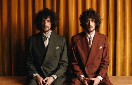 Twee mannen met krullend haar in vintage pakken, zittend voor een okergele gordijnachtergrond.