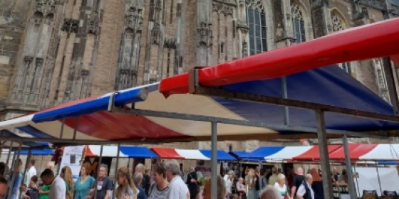 Marktkramen met rood-wit-blauwe zeilen en drukke menigte voor een grote oude kerk onder een bewolkte hemel.