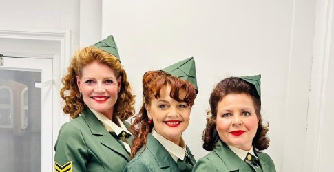 Drie vrouwen in groene vintage militaire uniformen staan naast elkaar in een lichte kamer en glimlachen.