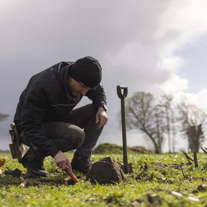Persoon met een metaaldetector en schop onderzoekt de grond op een veld onder een bewolkte hemel.