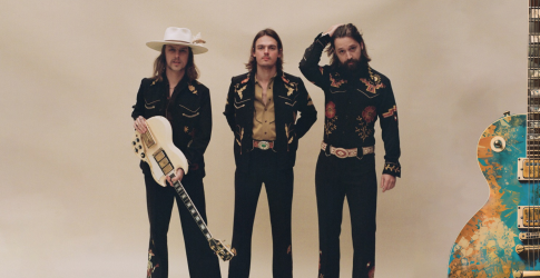 Drie mannen in westernkleding poseren met gitaren in een studio met een beige achtergrond en kleurrijke gitaar.