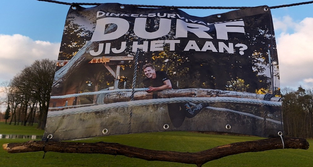 Spandoek met de tekst 'DINKEL SURVIVAL DURF JIJ HET AAN?' bij een outdoor survivalparcours.