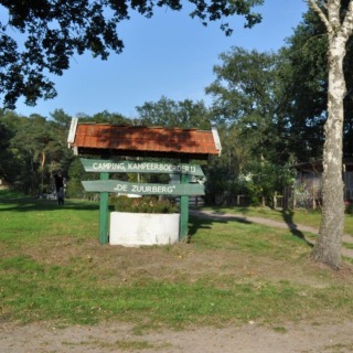 Entree van Camping Kampeerboerderij De Zuurberg met houten bord aan een zandpad en bomen op de achtergrond.