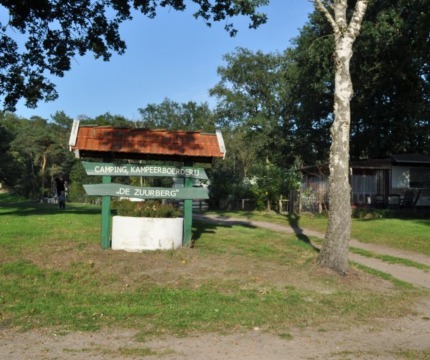 Entree van Camping Kampeerboerderij De Zuurberg met houten bord aan een zandpad en bomen op de achtergrond.
