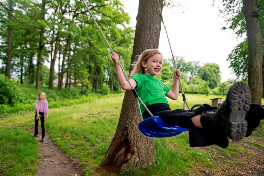 Twee meisjes spelen buiten in een bos; een meisje zit lachend op een schommel aan een boom.
