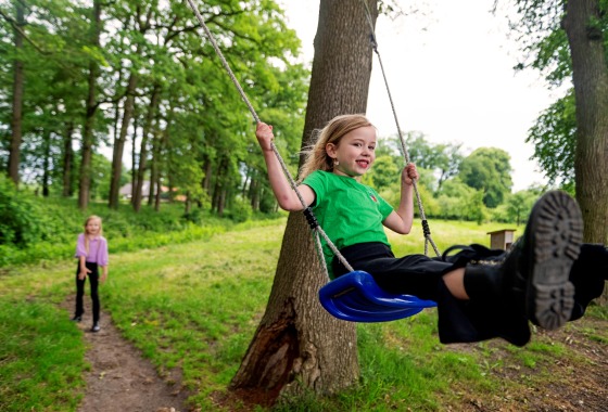 Twee meisjes spelen buiten in een bos; een meisje zit lachend op een schommel aan een boom.