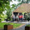 Groep mensen zit samen op een terras in een groene tuin, voor een huis met een rood pannendak.