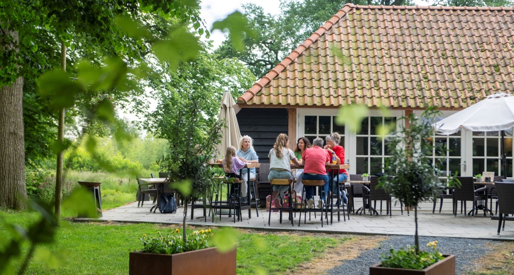 A group of people enjoying conversation on an outdoor patio in a lush garden near a tiled-roof house.