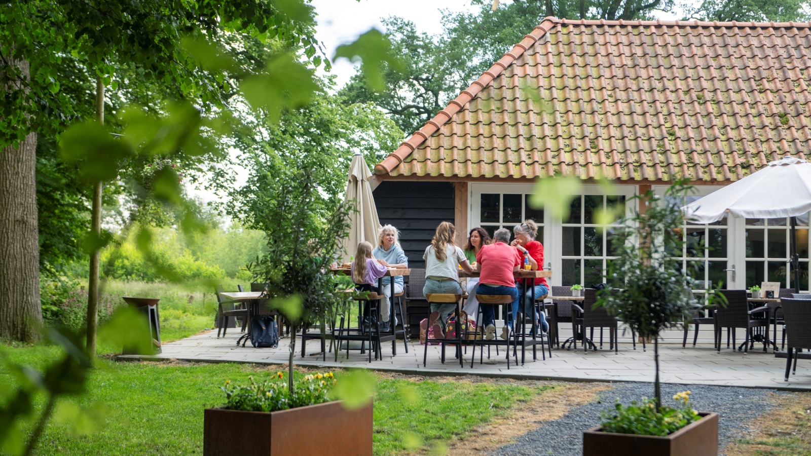 Groep mensen zit samen op een terras in een groene tuin, voor een huis met een rood pannendak.
