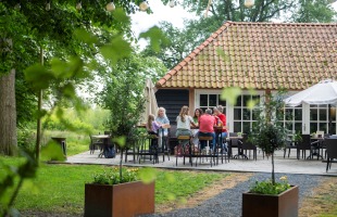 Groep mensen zit samen op een terras in een groene tuin, voor een huis met een rood pannendak.