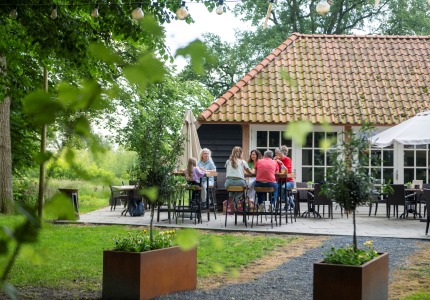 Groep mensen zit samen op een terras in een groene tuin, voor een huis met een rood pannendak.