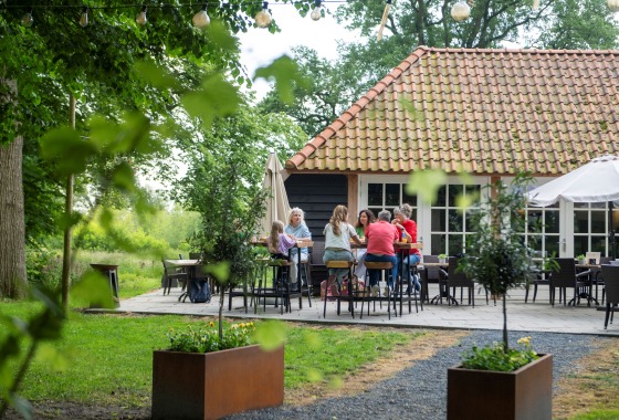 Groep mensen zit samen op een terras in een groene tuin, voor een huis met een rood pannendak.