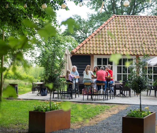 Groep mensen zit samen op een terras in een groene tuin, voor een huis met een rood pannendak.