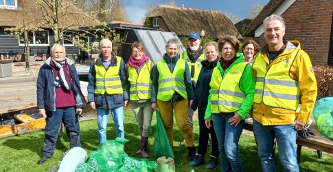 Groep vrijwilligers in gele hesjes poseren buiten met ingezameld afval in groene zakken op het gras.
