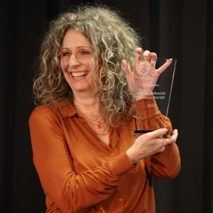 Vrouw met krullend haar in bruin shirt glimlacht en houdt een glazen awardtrofee op een podium vast.