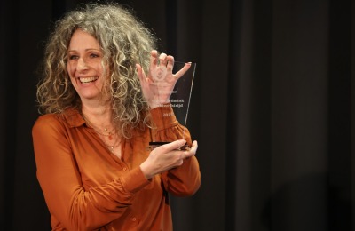 Vrouw met krullend haar in bruin shirt glimlacht en houdt een glazen awardtrofee op een podium vast.