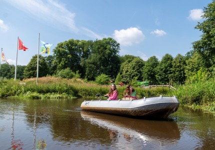 Twee vrouwen varen in een roeiboot op een rivier, met vlaggen en bomen op de achtergrond op een zonnige dag.