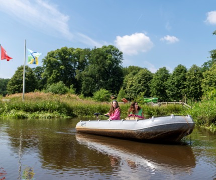 Twee vrouwen varen in een roeiboot op een rivier, met vlaggen en bomen op de achtergrond op een zonnige dag.