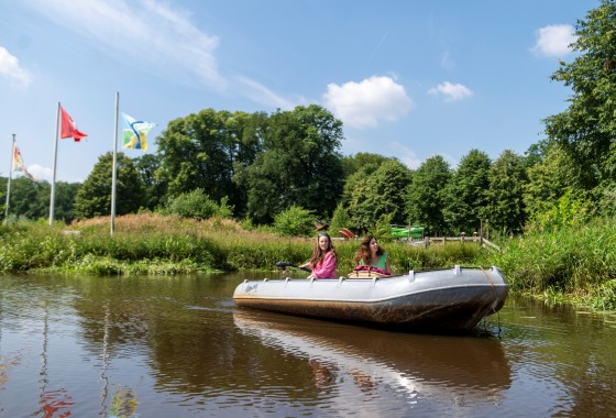 Twee vrouwen varen in een roeiboot op een rivier, met vlaggen en bomen op de achtergrond op een zonnige dag.
