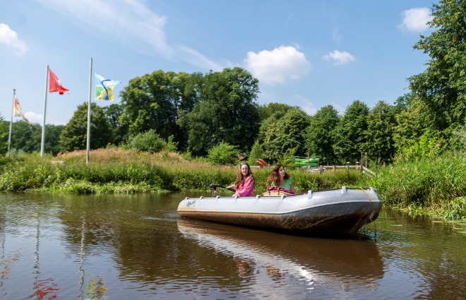 Zwei Frauen fahren in einem Ruderboot auf einem Fluss, dahinter Flaggen und Bäume an einem sonnigen Tag.
