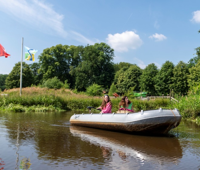 Twee vrouwen varen in een roeiboot op een rivier, met vlaggen en bomen op de achtergrond op een zonnige dag.