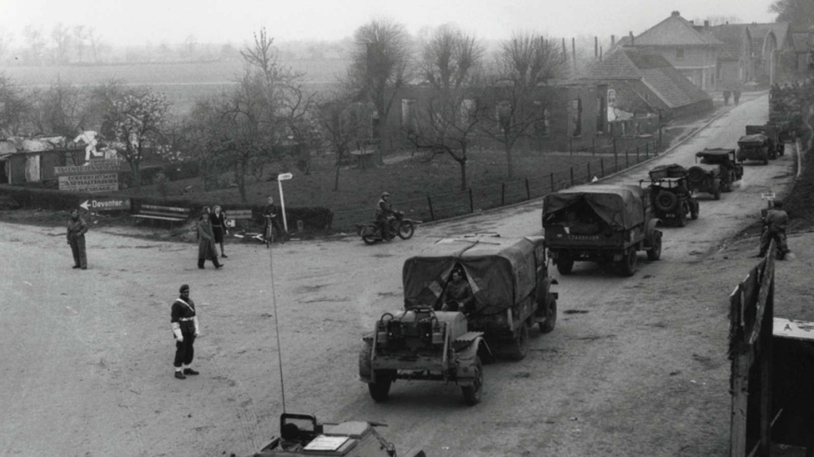Militairen en voertuigen passeren een kruispunt in een dorp, mogelijk tijdens de Tweede Wereldoorlog.
