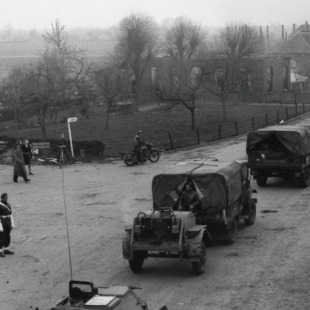 Militairen en voertuigen passeren een kruispunt in een dorp, mogelijk tijdens de Tweede Wereldoorlog.