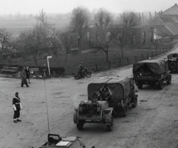 Militairen en voertuigen passeren een kruispunt in een dorp, mogelijk tijdens de Tweede Wereldoorlog.
