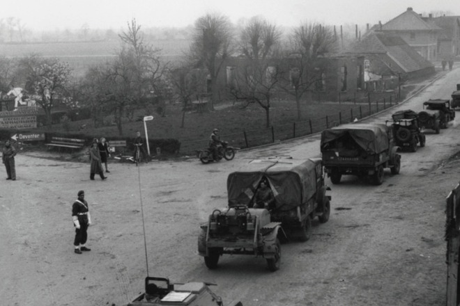 Militairen en voertuigen passeren een kruispunt in een dorp, mogelijk tijdens de Tweede Wereldoorlog.