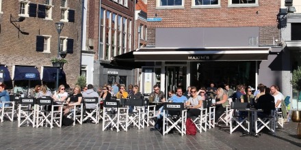 People enjoying a sunny day at an outdoor terrace of Maxim's café in a historic European city square.