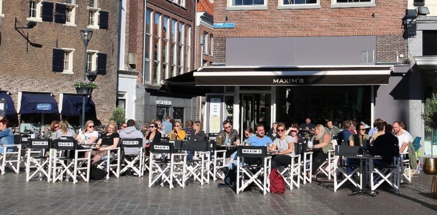 Mensen genieten van een zonnig terras bij Maxim's café in een historisch stadscentrum in Nederland.