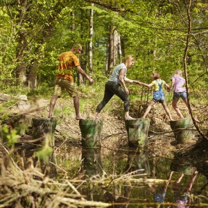 Een gezin steekt samen een beekje over op boomstammen in een zonnig bos, genietend van de natuur.