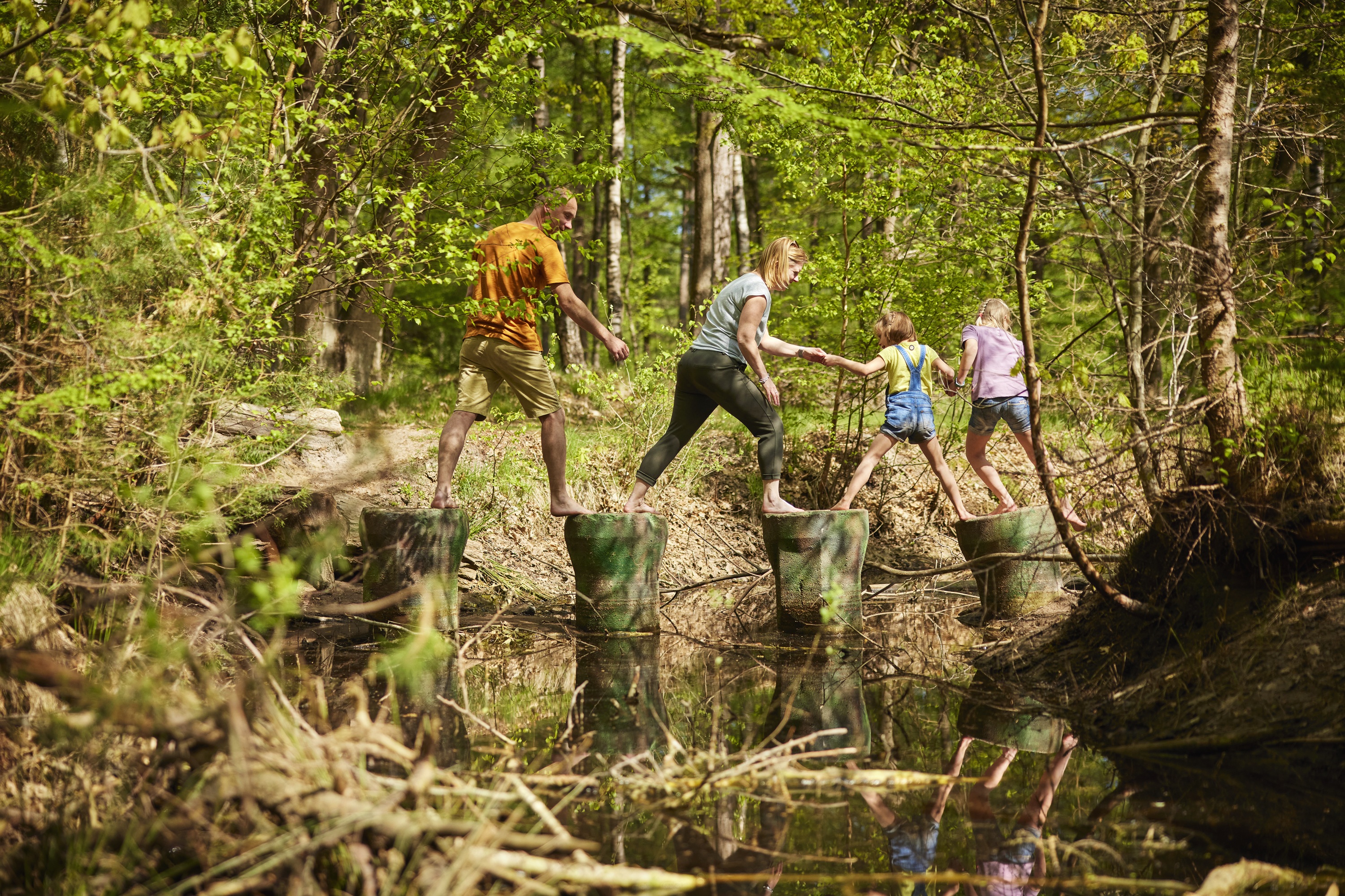 Een gezin steekt samen een beekje over op boomstammen in een zonnig bos, genietend van de natuur.