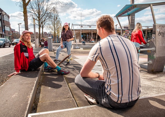 Drie jongeren zitten en staan bij een skateplek in de stad, terwijl ze met elkaar praten op een zonnige dag.