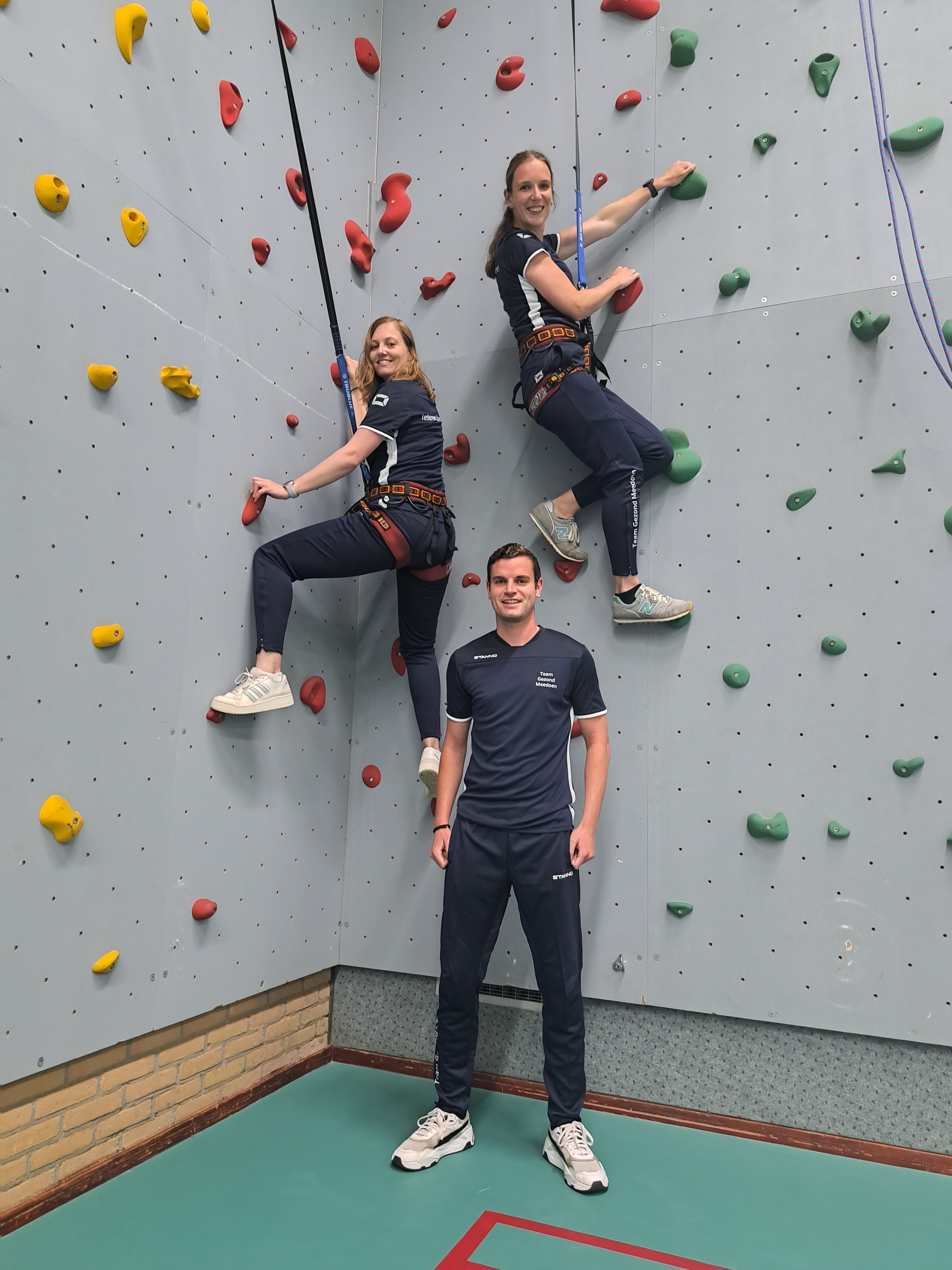 Twee vrouwen klimmen op een klimwand terwijl een man in sportkleding eronder op de grond staat en lacht.