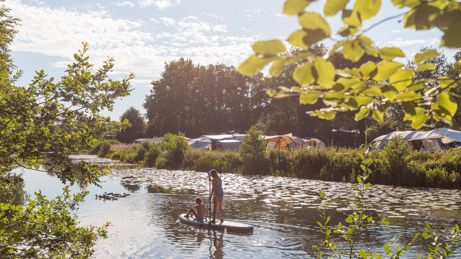 Twee mensen op een paddleboard op een rustige rivier, met tenten en bomen op de achtergrond tijdens de zomer.