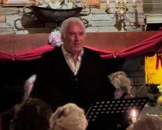 Een oudere man spreekt voor een klein publiek in een sfeervolle kamer met kaarslicht en decoratie.