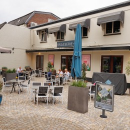 Buitenterras van een brasserie met stoelen, tafels en enkele etende gasten op een zonnige dag.